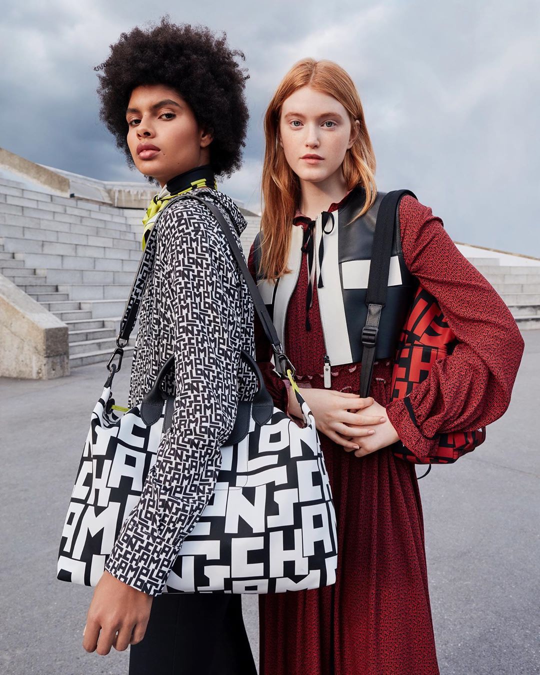 Two fashion models pose outdoors with Longchamp logo bags, one in a black and white pattern and one in red and black.