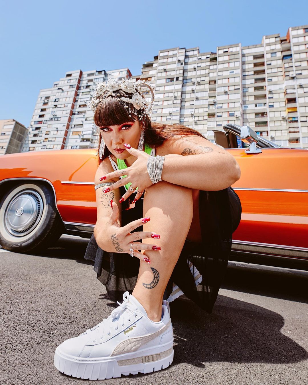 Woman in platform sneakers posing in front of an orange car and building with architectural details on a sunny day.