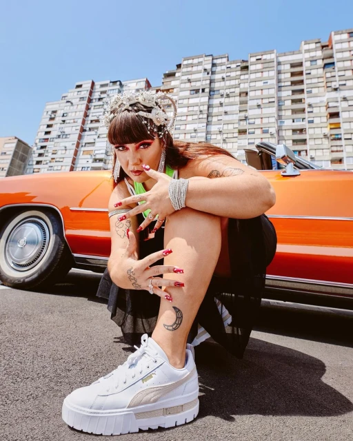 Woman in platform sneakers posing in front of an orange car and building with architectural details on a sunny day.