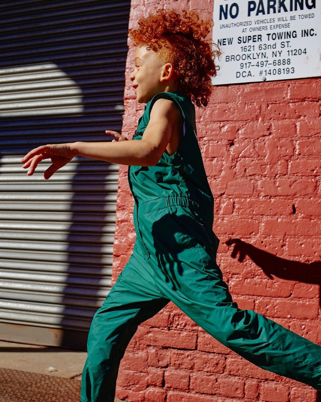 Child with red curly hair is outdoors, running past a red brick wall and gray metal roll-down door, wearing a green nylon sleeveless jumpsuit.