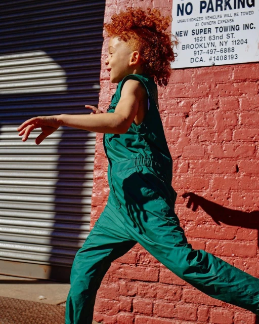 Child with red curly hair is outdoors, running past a red brick wall and gray metal roll-down door, wearing a green nylon sleeveless jumpsuit.