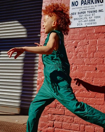 Child with red curly hair is outdoors, running past a red brick wall and gray metal roll-down door, wearing a green nylon sleeveless jumpsuit.