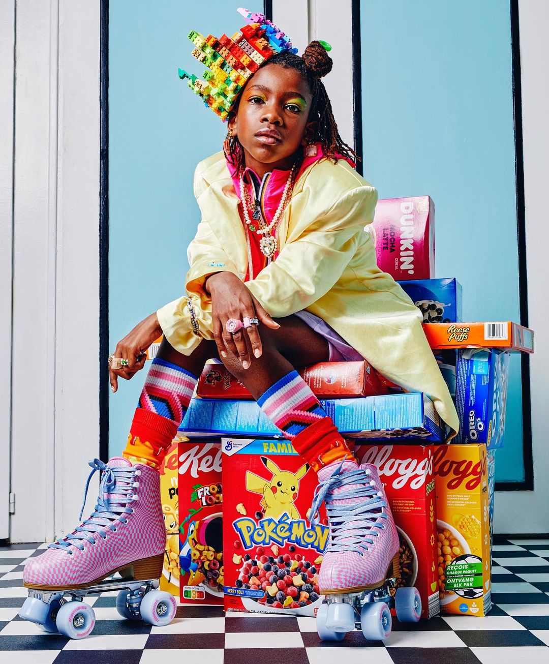 Young person in a yellow jacket and colorful crown, sitting on cereal boxes and wearing roller skates, posing in a studio with a blue background.