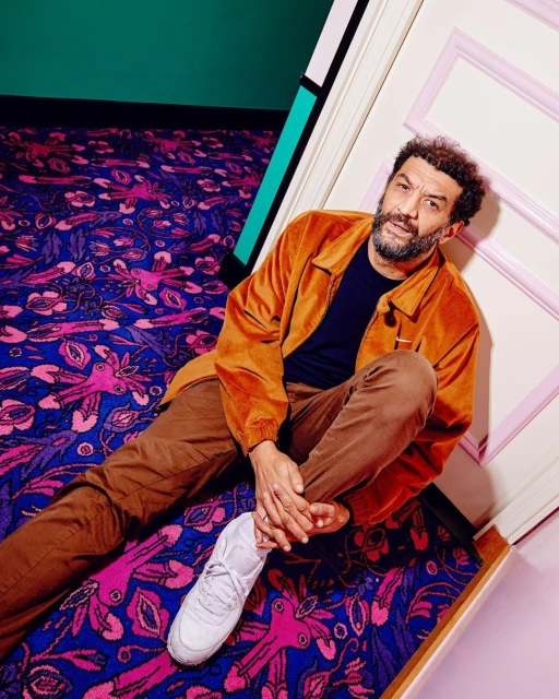 An adult man with a beard sits on a floral carpet, leaning against a wall, wearing a rust-colored jacket, brown pants, and white sneakers.