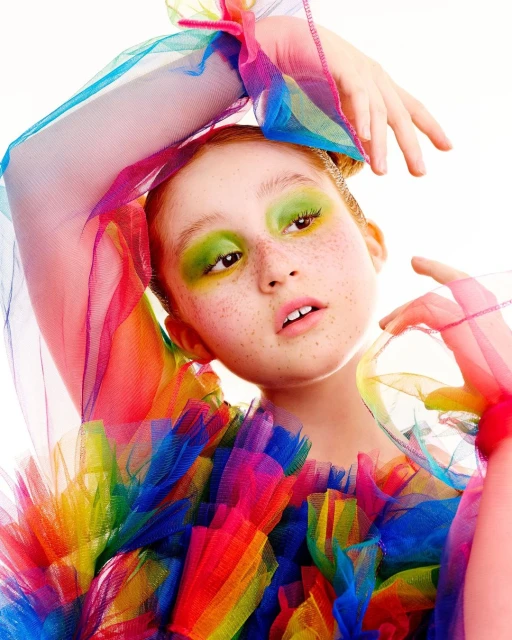 Close-up portrait of a young red-haired girl with freckles, wearing bright green eye shadow and a colorful tulle top in a photo studio.