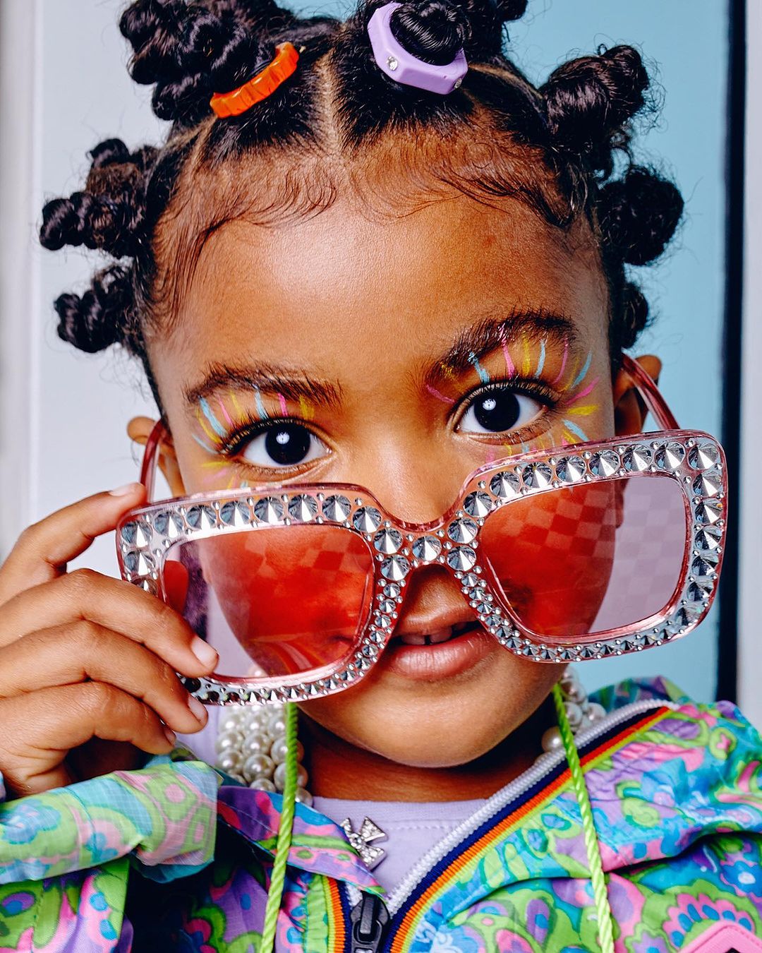 Close-up of a young child with colorful face makeup and hair accessories, wearing oversized, jeweled sunglasses and a patterned jacket.