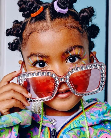 Close-up of a young child with colorful face makeup and hair accessories, wearing oversized, jeweled sunglasses and a patterned jacket.