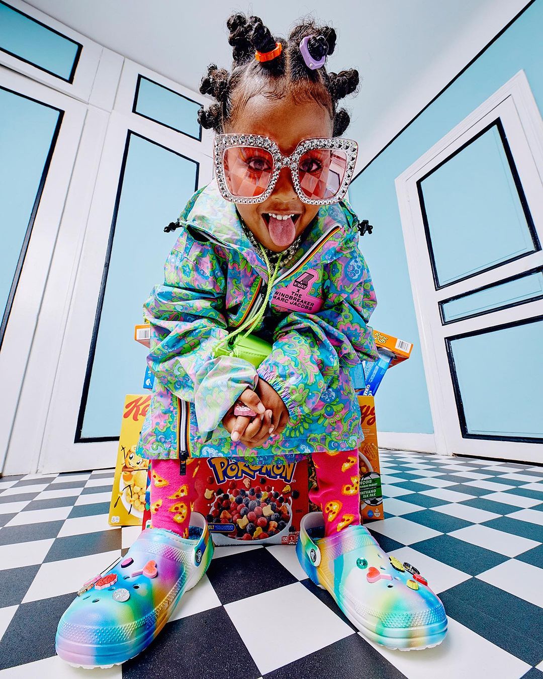 Cute child model wearing colorful clothing and oversized glasses poses with tongue out while seated on cereal boxes in photo studio.