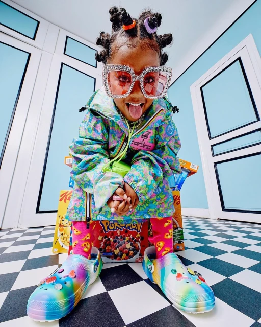 Cute child model wearing colorful clothing and oversized glasses poses with tongue out while seated on cereal boxes in photo studio.