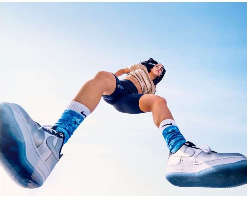 Low-angle view of a slender adult female with long dark hair and athletic wear, sporting white sneakers and blue socks, against a light blue sky.