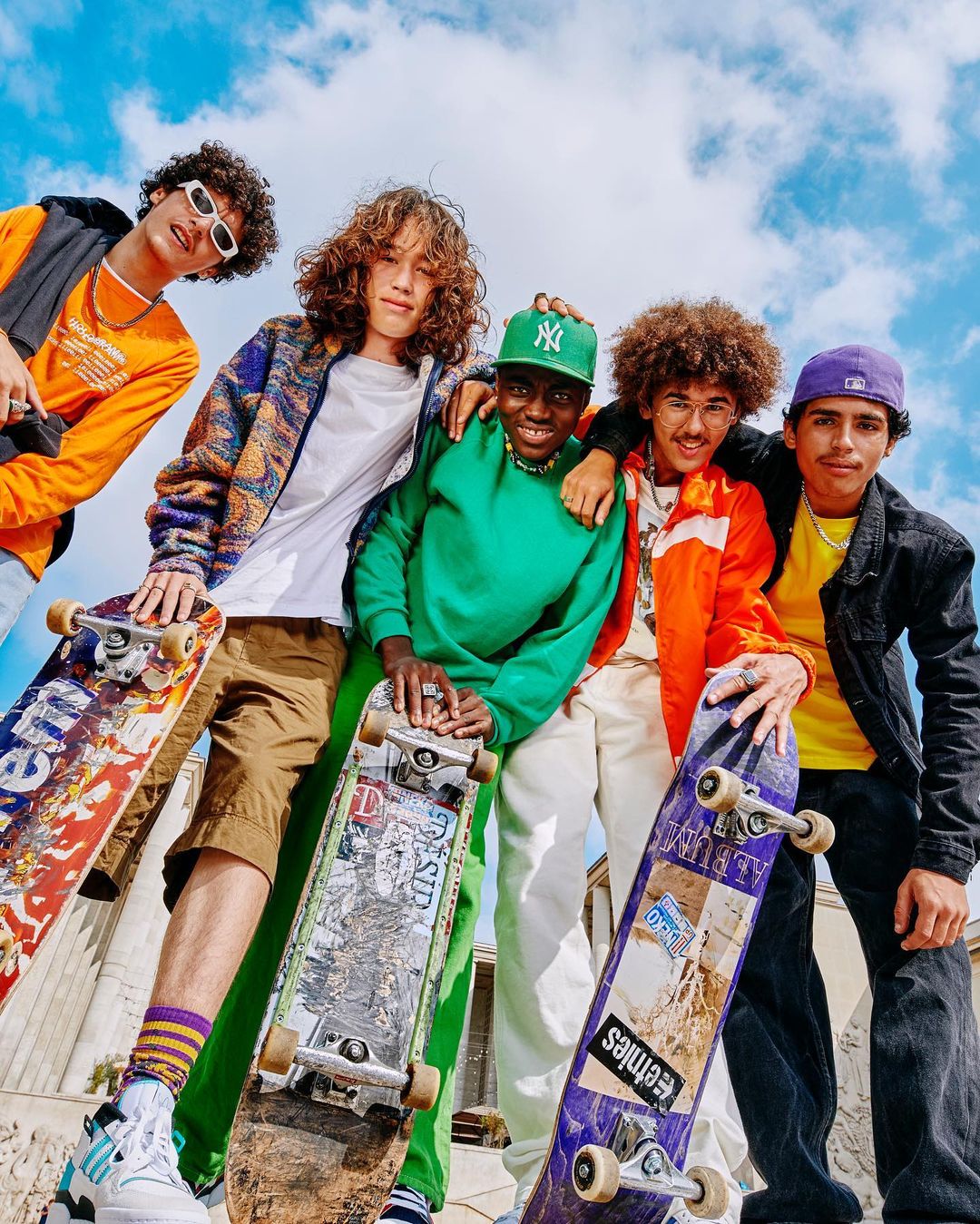 Five young men pose with skateboards against a blue sky. The group wears casual clothing and hats, creating a youthful, urban vibe.