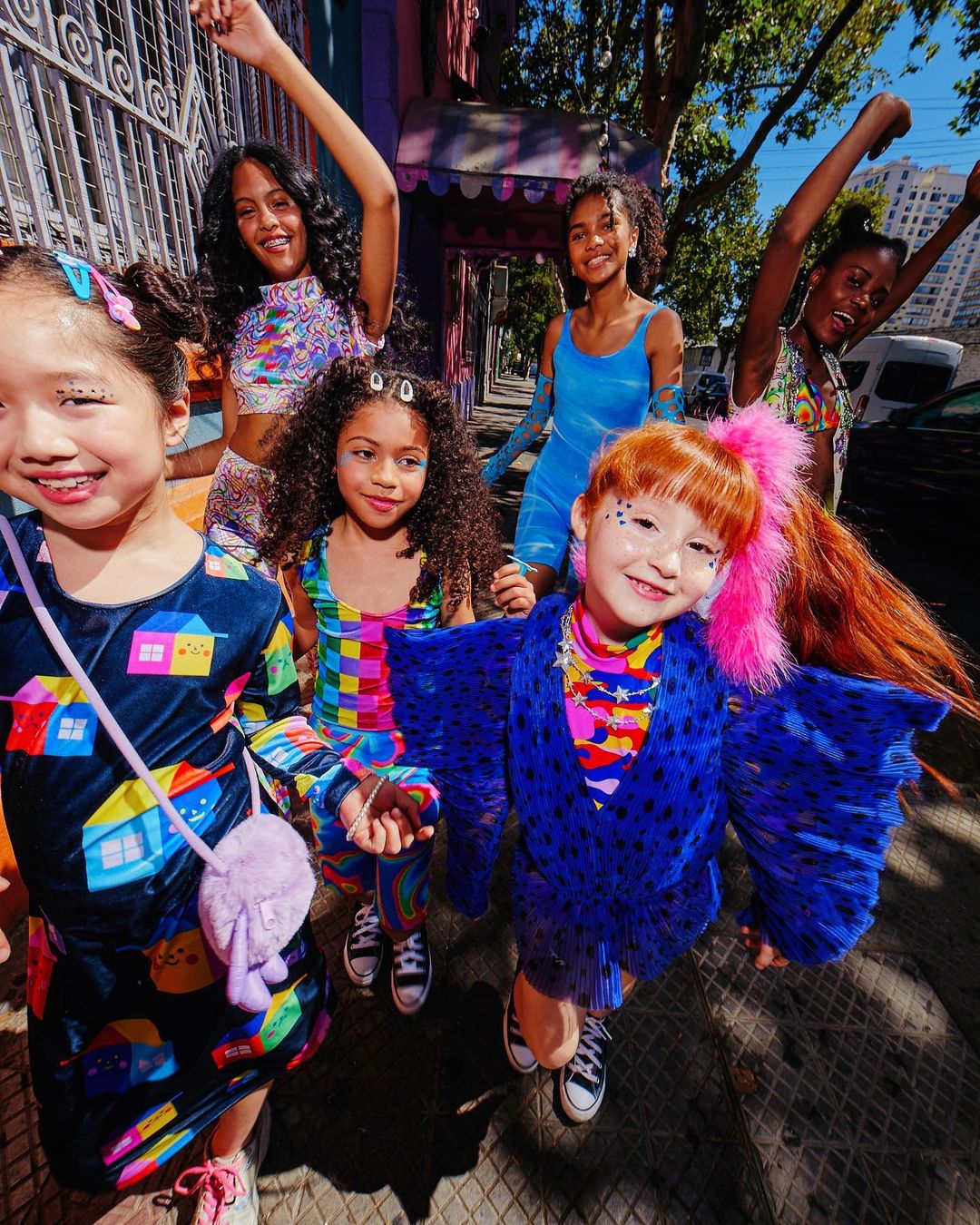 Five young girls in colorful clothing pose on a sidewalk in an urban area with buildings in the background on a bright day.