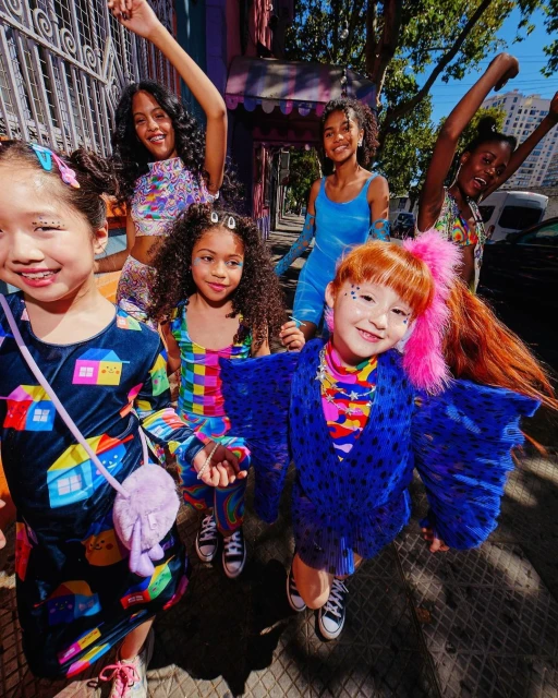 Five young girls in colorful clothing pose on a sidewalk in an urban area with buildings in the background on a bright day.