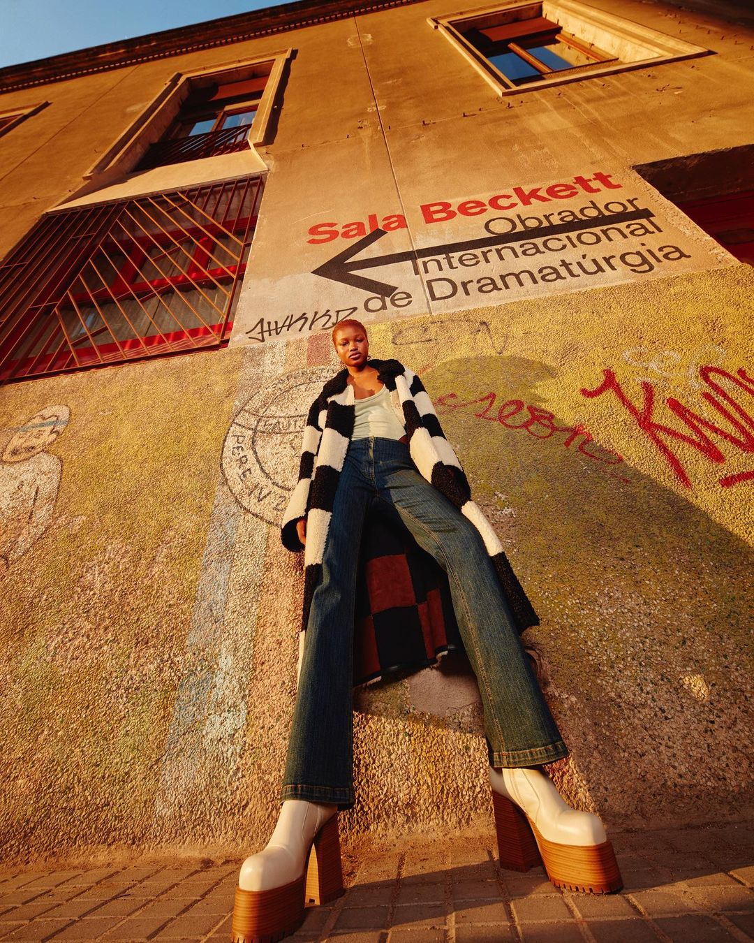 Woman poses in tall platform boots and a long coat against a wall with graffiti and theatre signage in an urban environment.