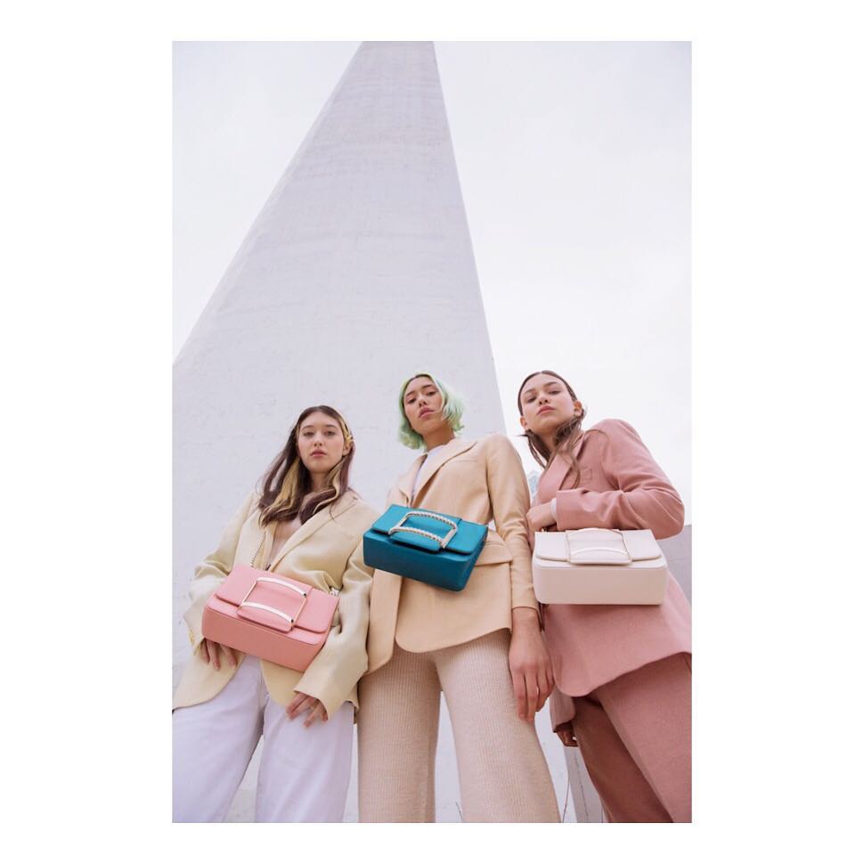 Three women pose with colorful handbags in front of a white pyramid structure, wearing stylish suits and matching monochrome attire.
