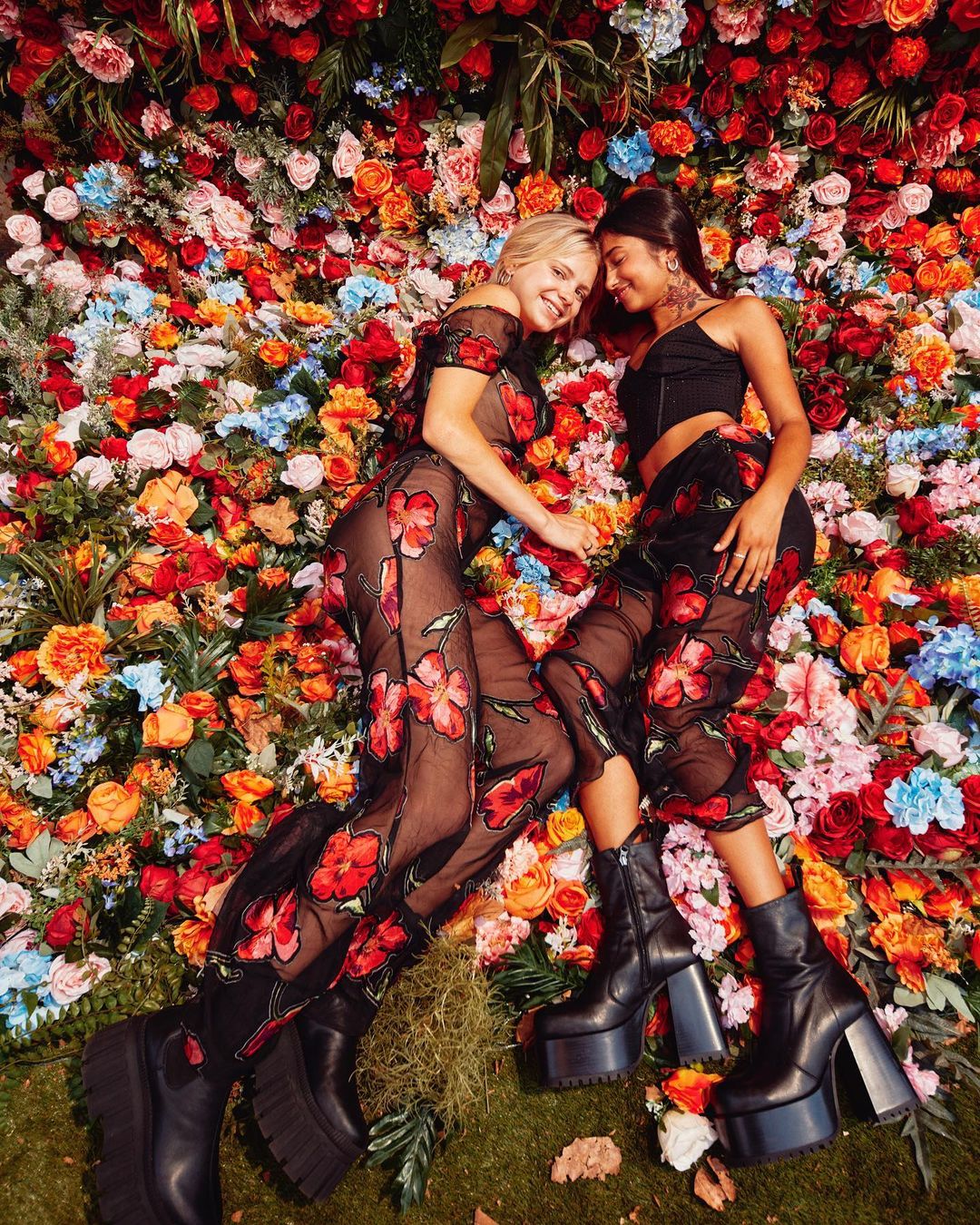 Two women lying side by side against a dense wall of colourful floral decorations, wearing black floral outfits and platform boots.