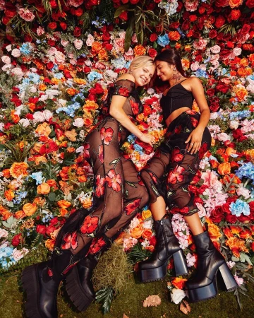 Two women lying side by side against a dense wall of colourful floral decorations, wearing black floral outfits and platform boots.