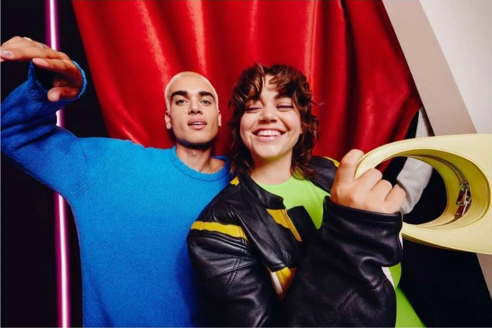 Two smiling people in a studio. One with short blonde hair wears a blue sweater, the other wears a green top and leather jacket.