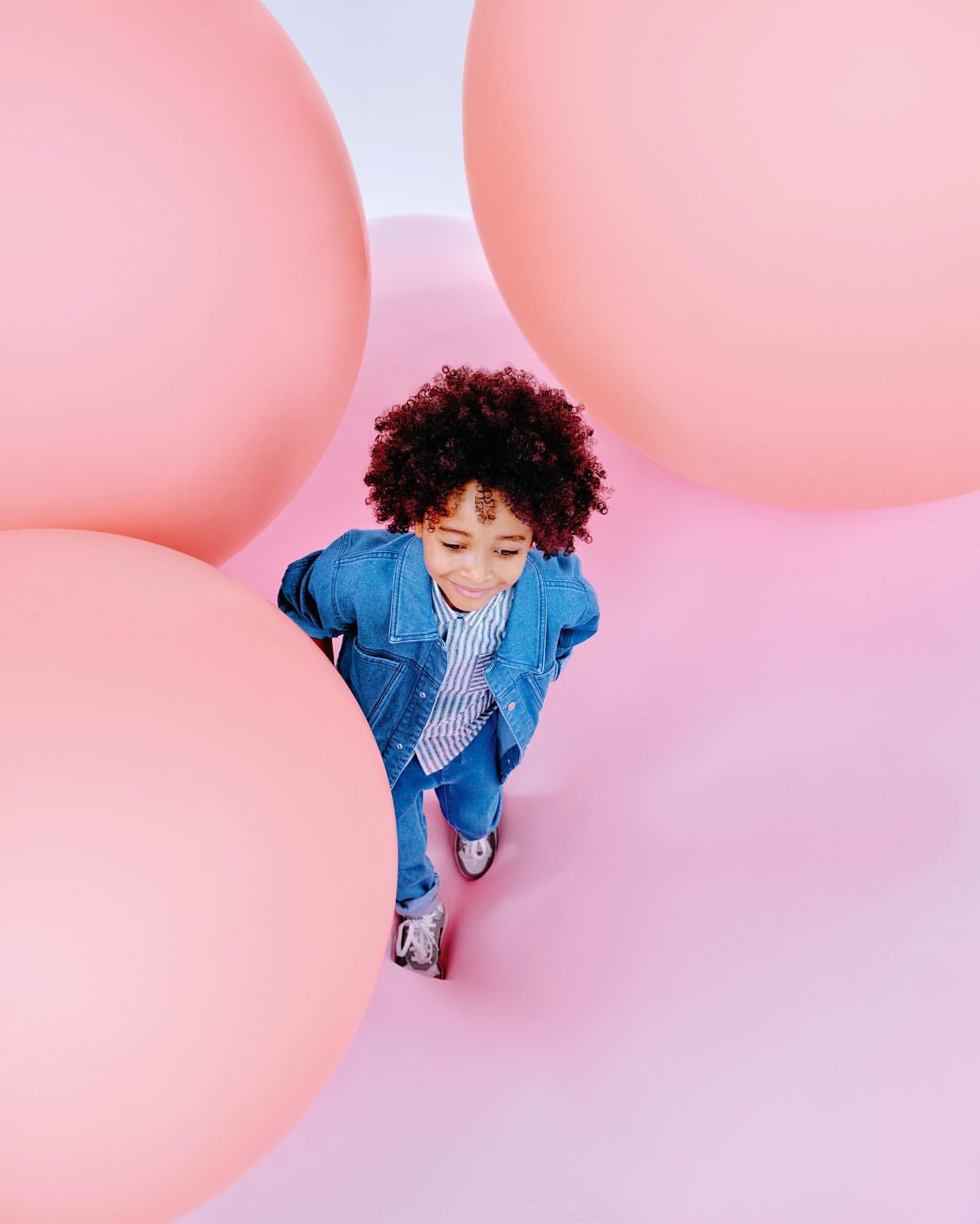 A child with curly hair stands surrounded by large pink spheres, wearing a denim jacket, striped shirt, and jeans on a pink surface.