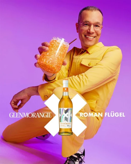 Man in yellow jumpsuit poses with a cocktail drink and a bottle of Glenmorangie whisky against a purple background.