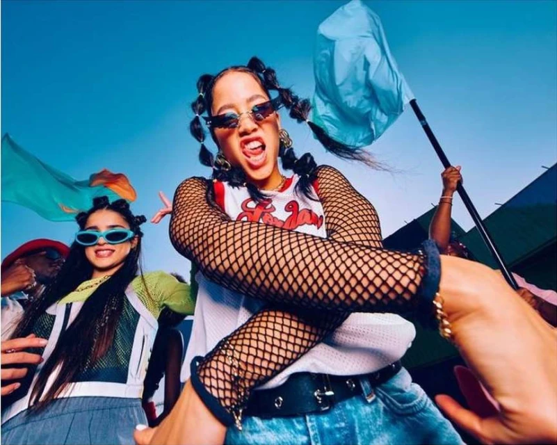 Group of young people in sunglasses and casual wear outdoors with blue sky and flags, expressing carefree and youthful energy.