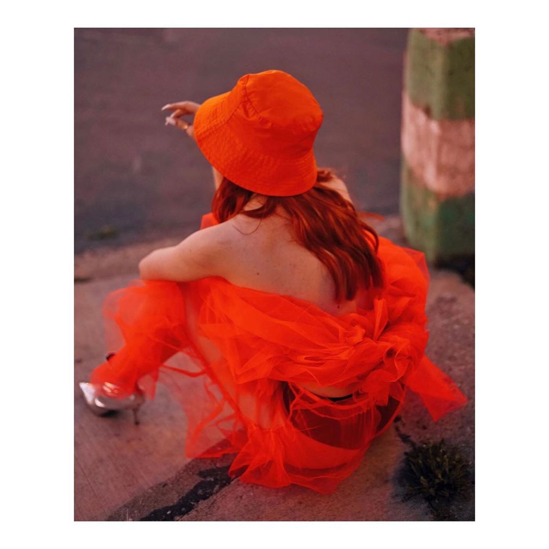 Back view of an adult female wearing a bright orange bucket hat and orange dress sitting on concrete with metallic high heel shoes.