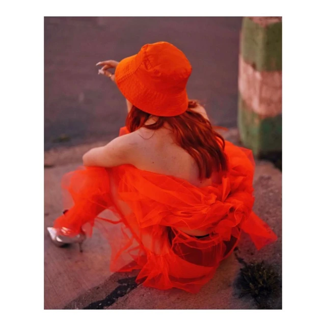 Back view of an adult female wearing a bright orange bucket hat and orange dress sitting on concrete with metallic high heel shoes.
