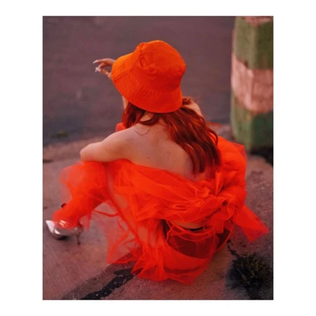 Back view of an adult female wearing a bright orange bucket hat and orange dress sitting on concrete with metallic high heel shoes.