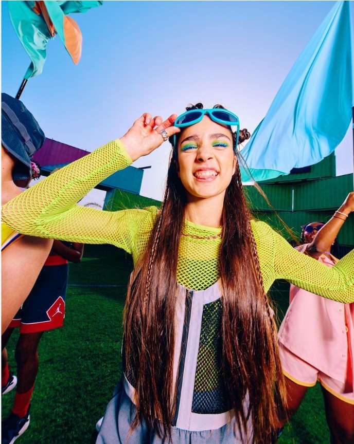 Smiling woman with braided brown hair, wearing blue sunglasses and a chartreuse net shirt, poses outdoors with a group on a sunny day.