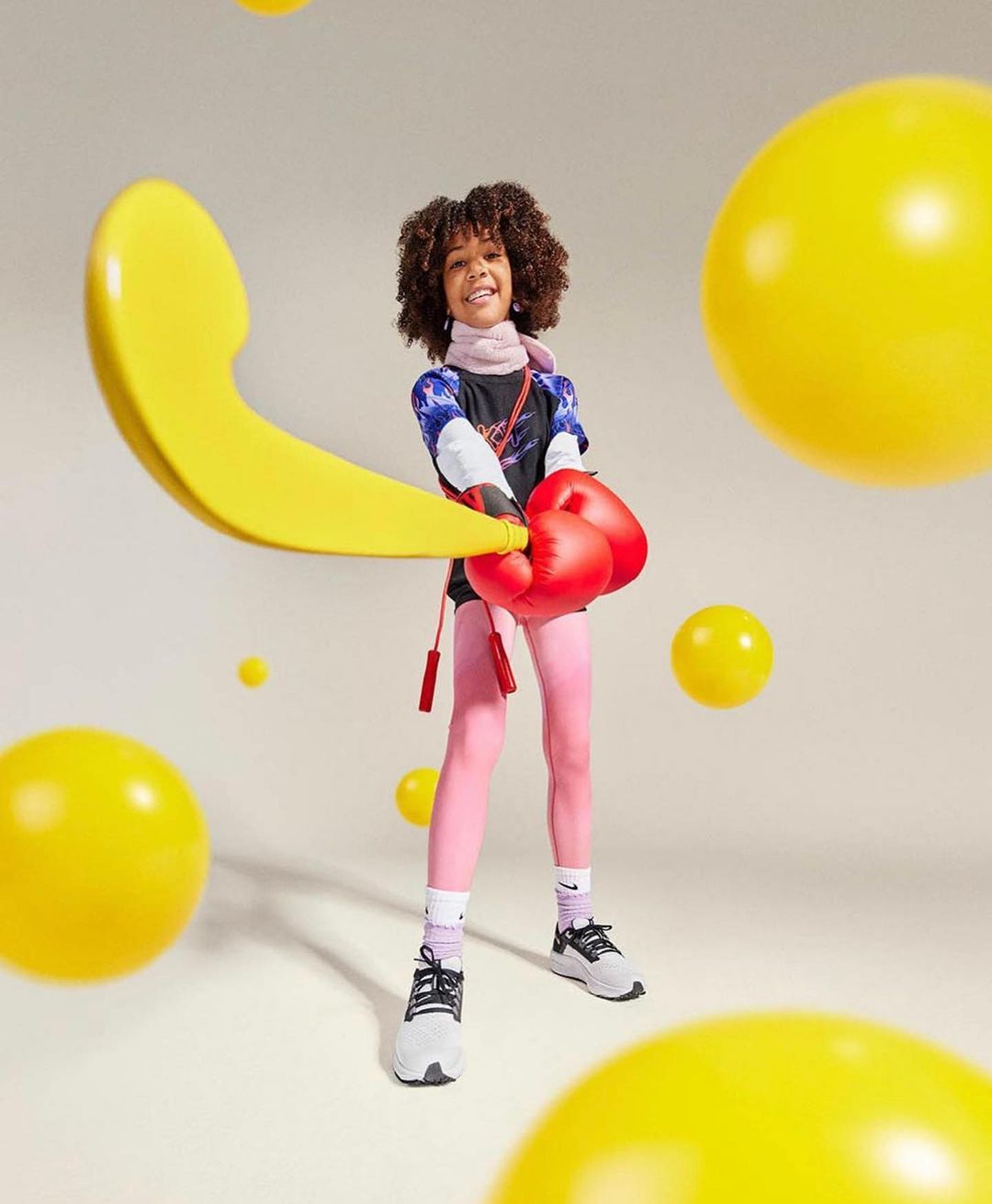 A young girl with curly hair is wearing boxing gloves, pink leggings, and sneakers, posing in a cream-colored studio with yellow spheres.