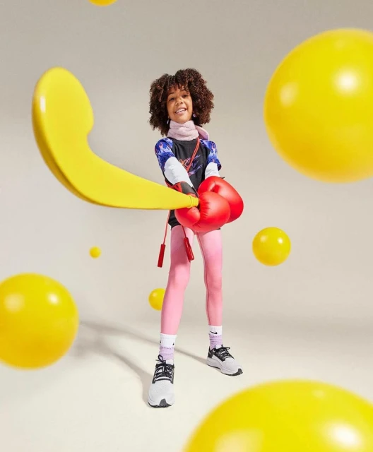 A young girl with curly hair is wearing boxing gloves, pink leggings, and sneakers, posing in a cream-colored studio with yellow spheres.