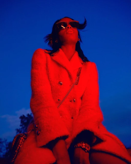 A person with sunglasses and an open coat poses in front of a blue sky in a red-tinted portrait. The subject wears jewelry and looks up.