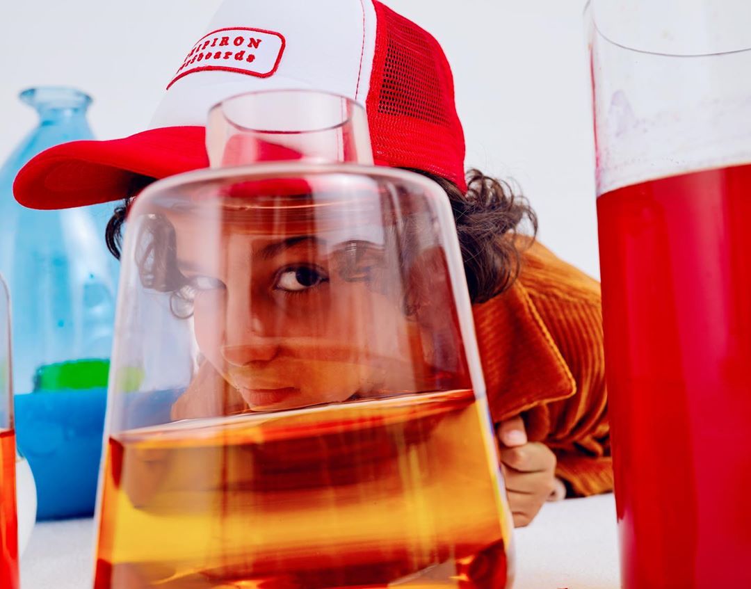 Child looks through glass beaker with red liquid in photo studio. They wear a baseball cap and a corduroy jacket with a white background.