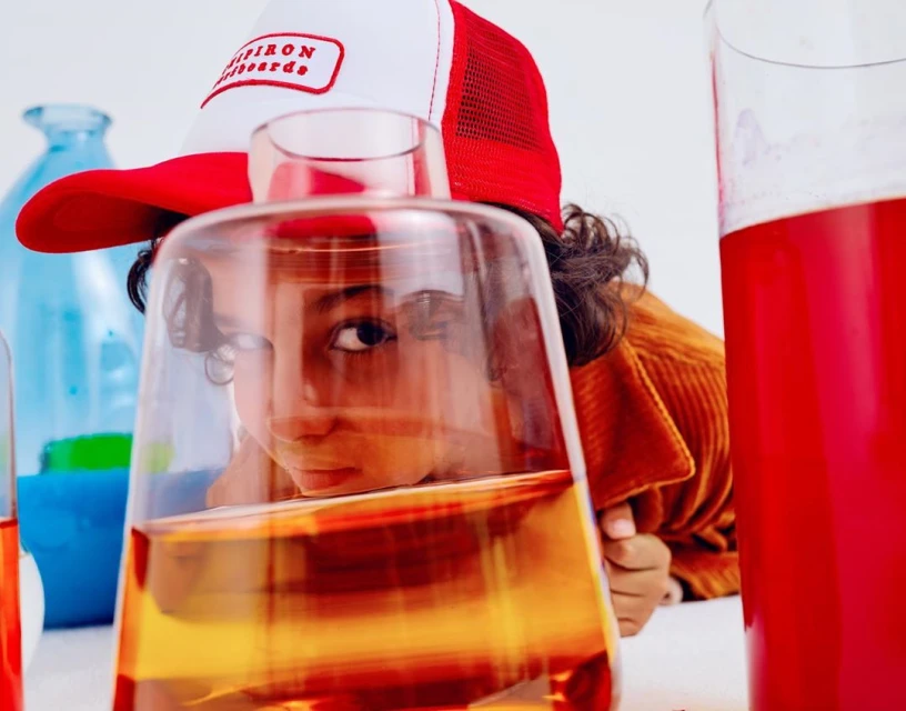 Child looks through glass beaker with red liquid in photo studio. They wear a baseball cap and a corduroy jacket with a white background.