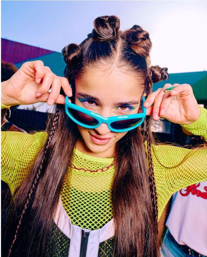 Portrait of a young woman with space buns, turquoise sunglasses and eyeshadow, wearing a mesh top and chain necklace, looking at camera.