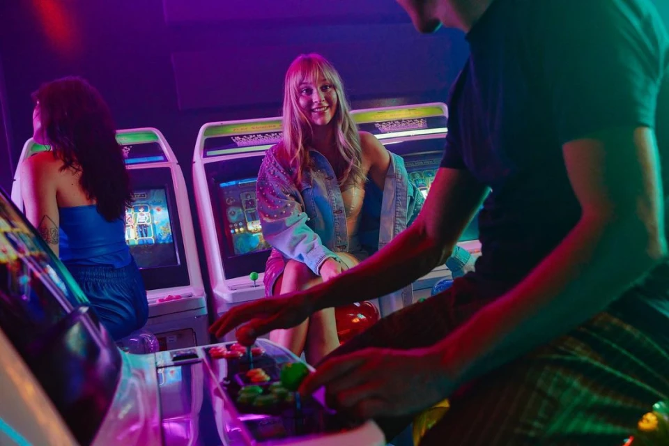 Three people are at an arcade, two are playing vintage arcade games while the third is looking on with a smile in a vivid setting.