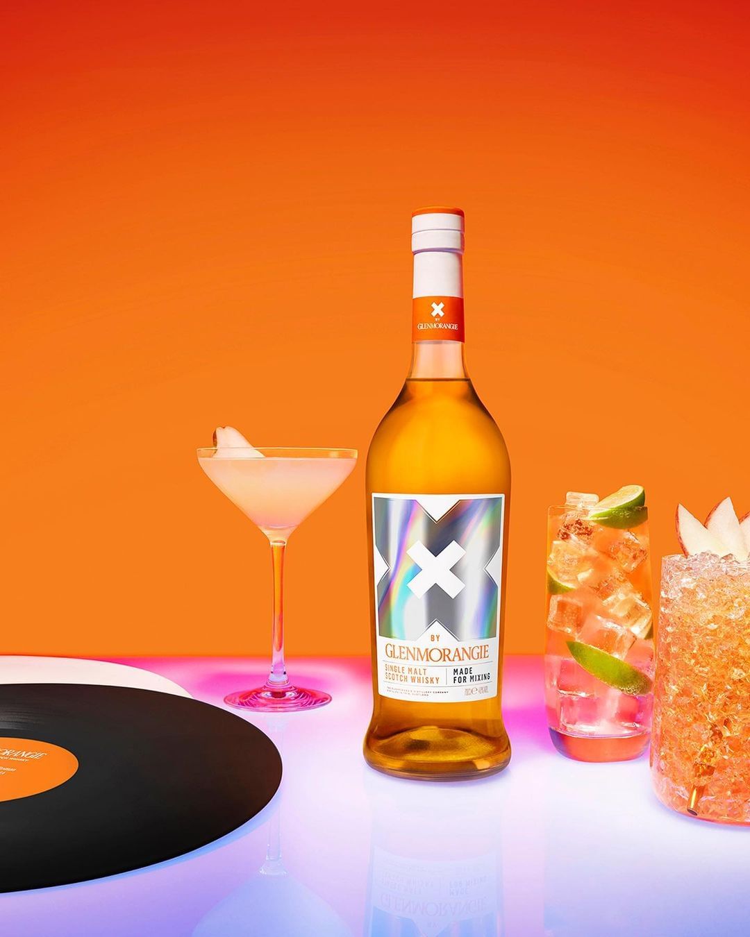 Glenmorangie scotch whisky bottle with two cocktails, record, against orange background, lit with neon pink and blue lights.