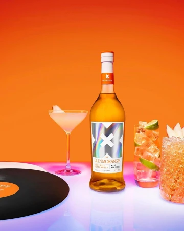 Glenmorangie scotch whisky bottle with two cocktails, record, against orange background, lit with neon pink and blue lights.