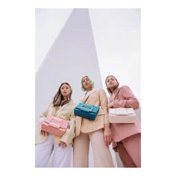 Three female models in pastel blazers holding colorful handbags against a tall concrete structure outdoors.