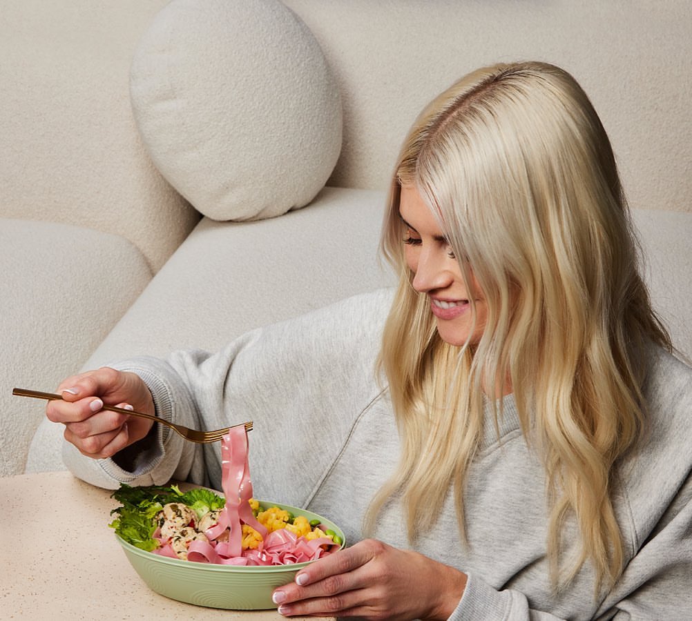 Woman eats a vibrant salad with pink noodles at a light-toned table, dressed in a grey sweatshirt, with a bright, happy expression.