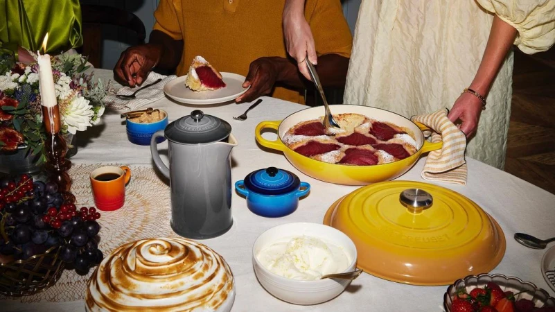 Two people at a table with a pear dessert, coffee, flowers, and Le Creuset cookware in a warm, inviting indoor setting.