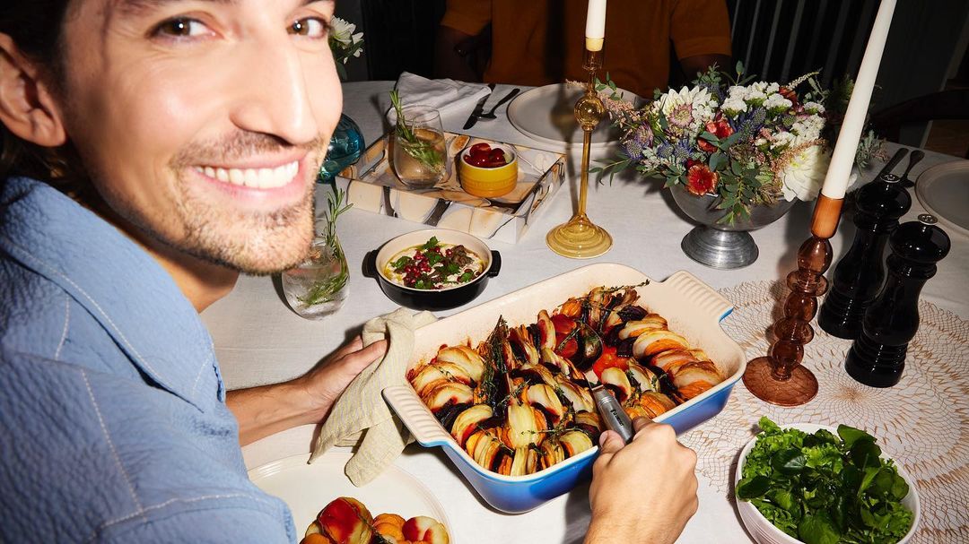 Smiling man with baked vegetables at dining table. Enjoying a meal at home with fresh salad, flowers, candles, and beverages.