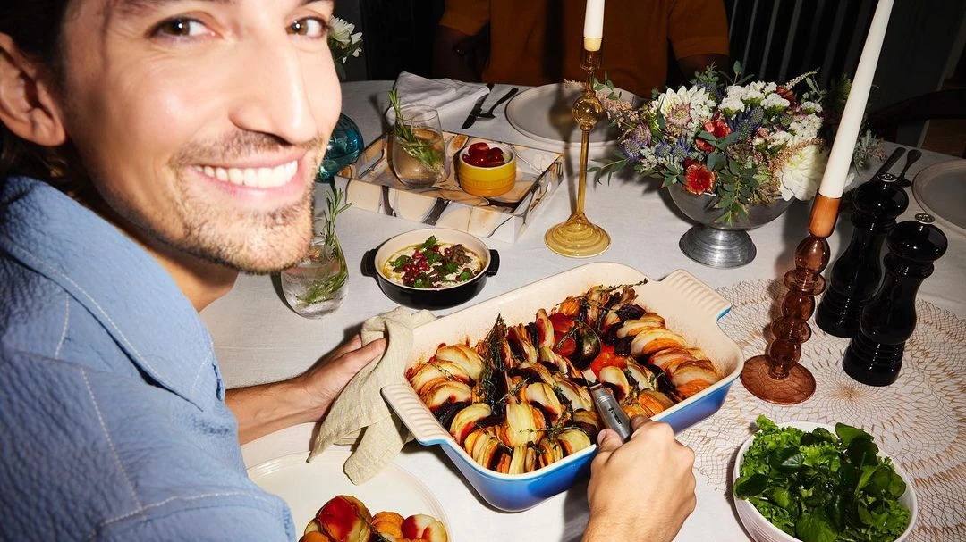 Smiling man with baked vegetables at dining table. Enjoying a meal at home with fresh salad, flowers, candles, and beverages.