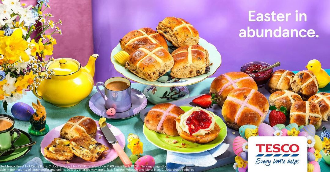 Festive Easter still life featuring hot cross buns, tea, flowers, and Easter decorations on a table against a purple background.