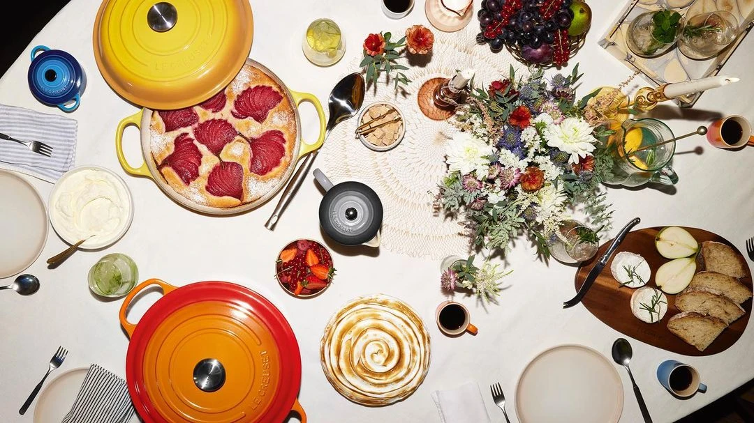 Overhead shot of an elegantly set table with Le Creuset cookware, pastries, and fresh fruit. A colorful floral centerpiece adds a touch of freshness.