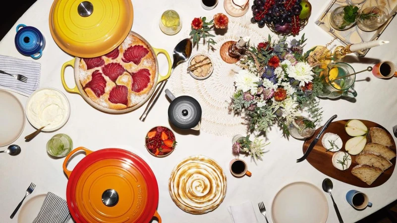 Overhead shot of an elegantly set table with Le Creuset cookware, pastries, and fresh fruit. A colorful floral centerpiece adds a touch of freshness.