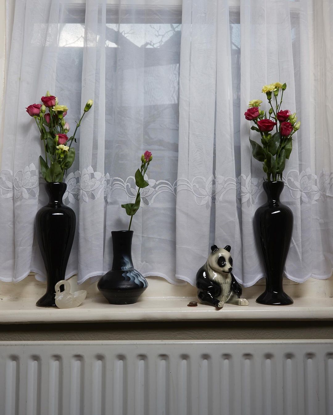 Still life of vases with roses and a panda figurine arranged on a windowsill in front of sheer white curtains and a radiator.