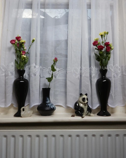 Still life of vases with roses and a panda figurine arranged on a windowsill in front of sheer white curtains and a radiator.