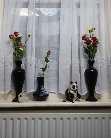 Still life of vases with roses and a panda figurine arranged on a windowsill in front of sheer white curtains and a radiator.