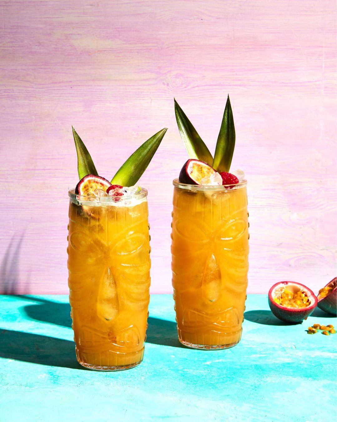 Two tropical cocktails in tiki glasses garnished with passion fruit and pineapple leaves, set against a vibrant pink and blue background.